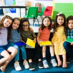 Kindergarten student friends using their notebooks and smiling while posing together in the school classroom.