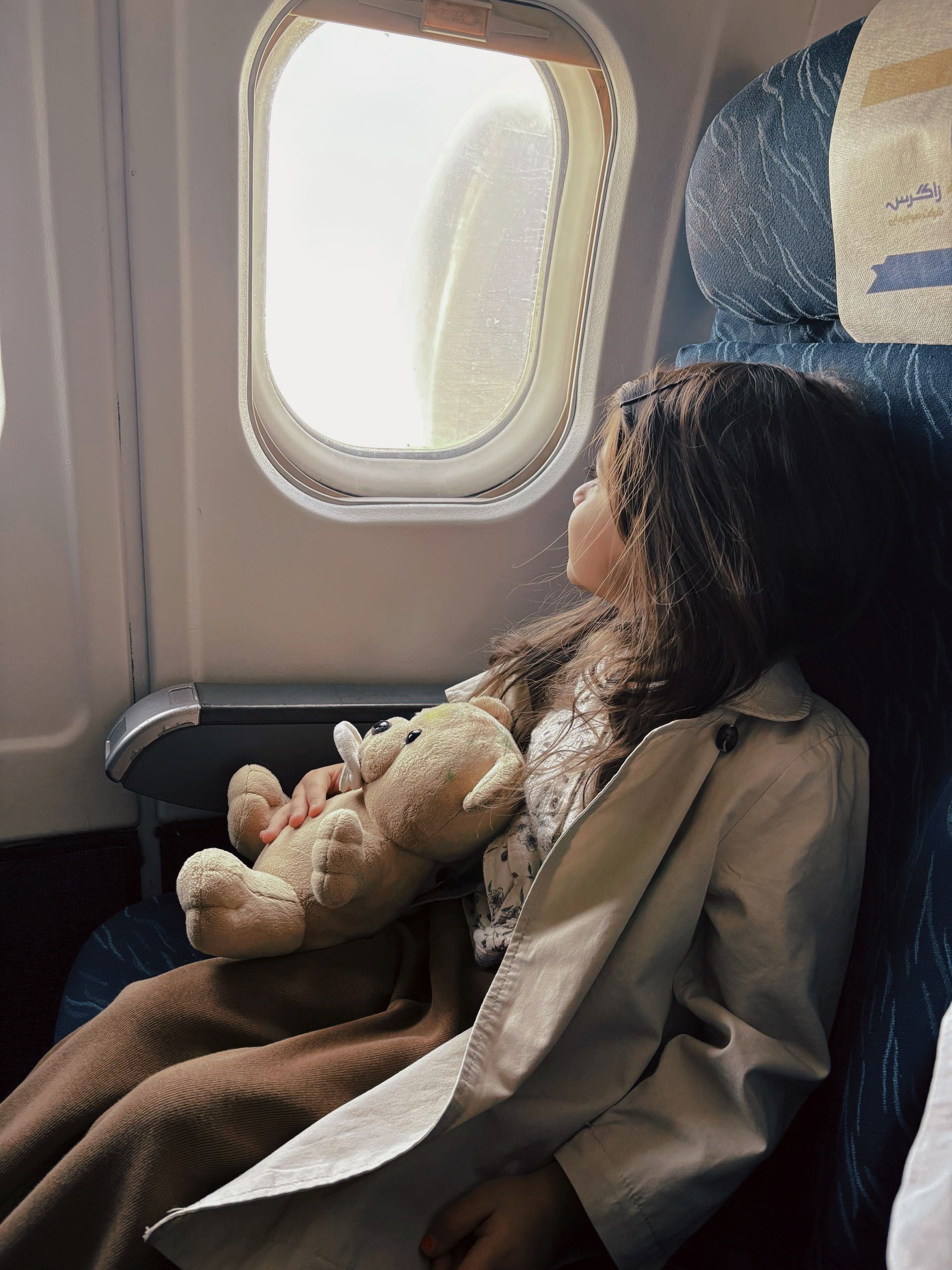 A young girl holding a teddy bear looks out an airplane window, flying peacefully.